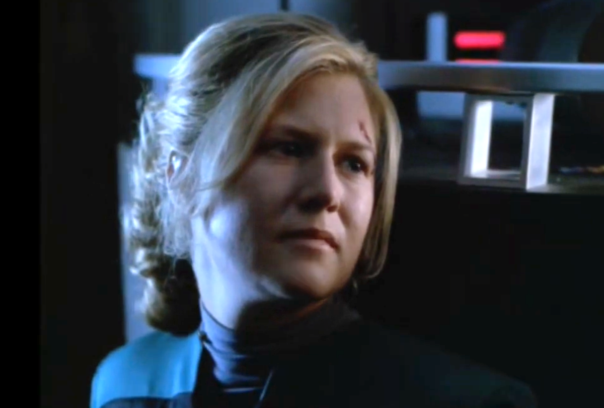 A close up of Ensign Samantha Wildman, a blonde woman in a teal Starfleet uniform, her hair pulled back, with a small cut on her forehead. She looks off to the side with a tense, concerned expression while standing in a dimly lit starship interior, metallic equipment and a red indicator light visible behind her. From the Star Trek: Voyager episode titled "Once Upon a Time," directed by John Kretchmer. First aired in 1998. Wildman is played by Nancy Hower.