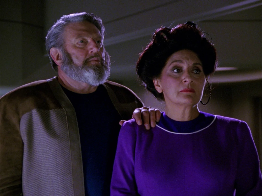 A medium shot of two older characters from Star Trek: The Next Generation, Sergey and Helena Rozhenko (Worf’s human foster parents). Sergey, a man with a thick grey beard and hair, wears a brown and tan textured jacket over a dark shirt. Helena stands beside him in a vibrant purple long-sleeved dress with white trim at the collar, her dark hair styled in an elegant updo. They both look off-camera with concerned, supportive expressions.